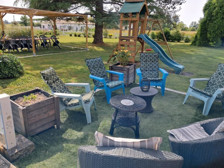 Espace détente extérieur avec transats, fauteuils colorés, table basse et aire de jeux pour enfants, entouré de bacs à plantes et de pelouse verte au gîte Les P'tites Cagouilles
